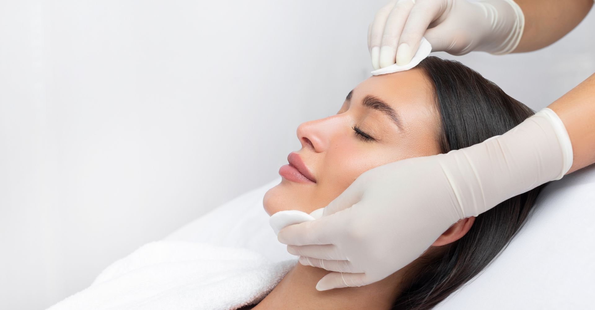 Mulher deitada em uma maca com rosto descansado enquanto uma esteticista passa algodão em sua pele para a limpeza de pele profunda.