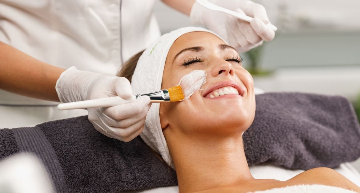 Mulher sorridente em maca enquanto uma esteticista passa máscara branca em seu rosto.