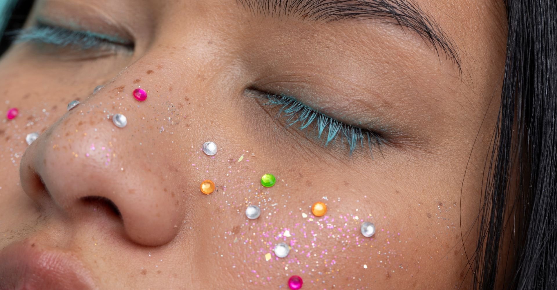 Close-up do rosto de uma pessoa com os olhos fechados, exibindo uma maquiagem criativa com cílios azuis vibrantes, glitter rosa e prateado, e pedrinhas coloridas em tons de rosa, laranja, verde e prata espalhadas pelo nariz e bochechas.