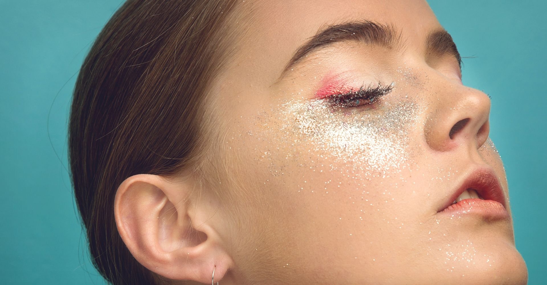 Close-up do perfil de uma pessoa com os olhos fechados, exibindo uma maquiagem artística com glitter prateado e rosa vívido ao redor dos olhos e bochechas.