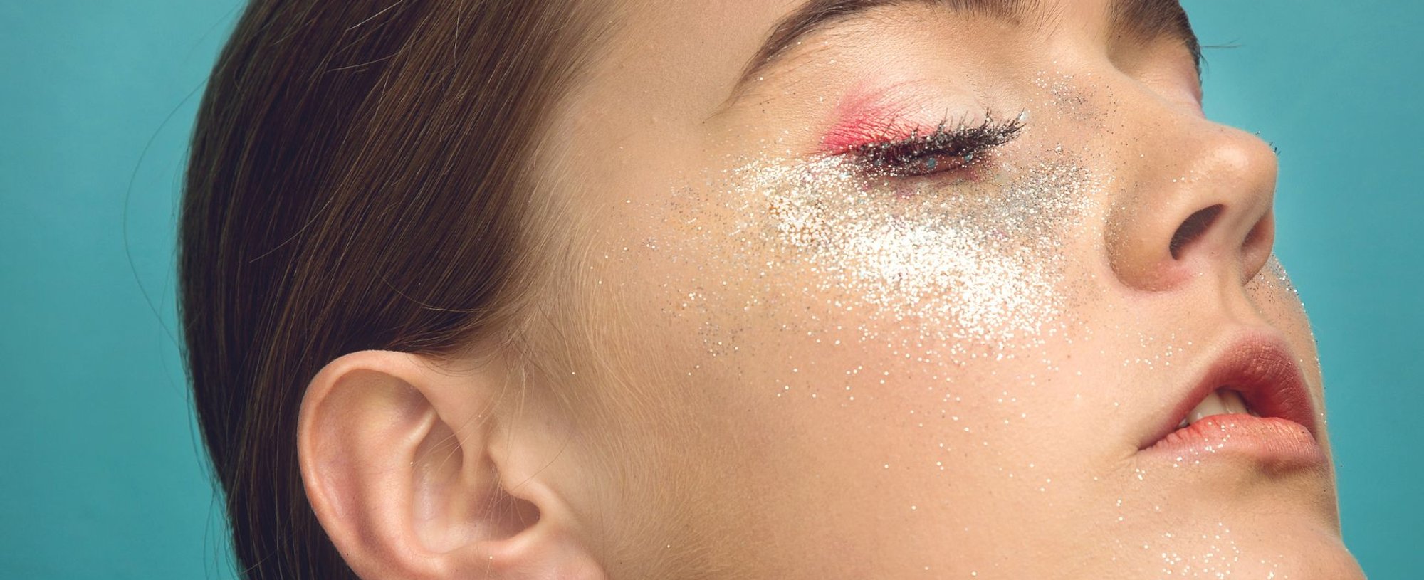 Close-up do perfil de uma pessoa com os olhos fechados, exibindo uma maquiagem artística com glitter prateado e rosa vívido ao redor dos olhos e bochechas.