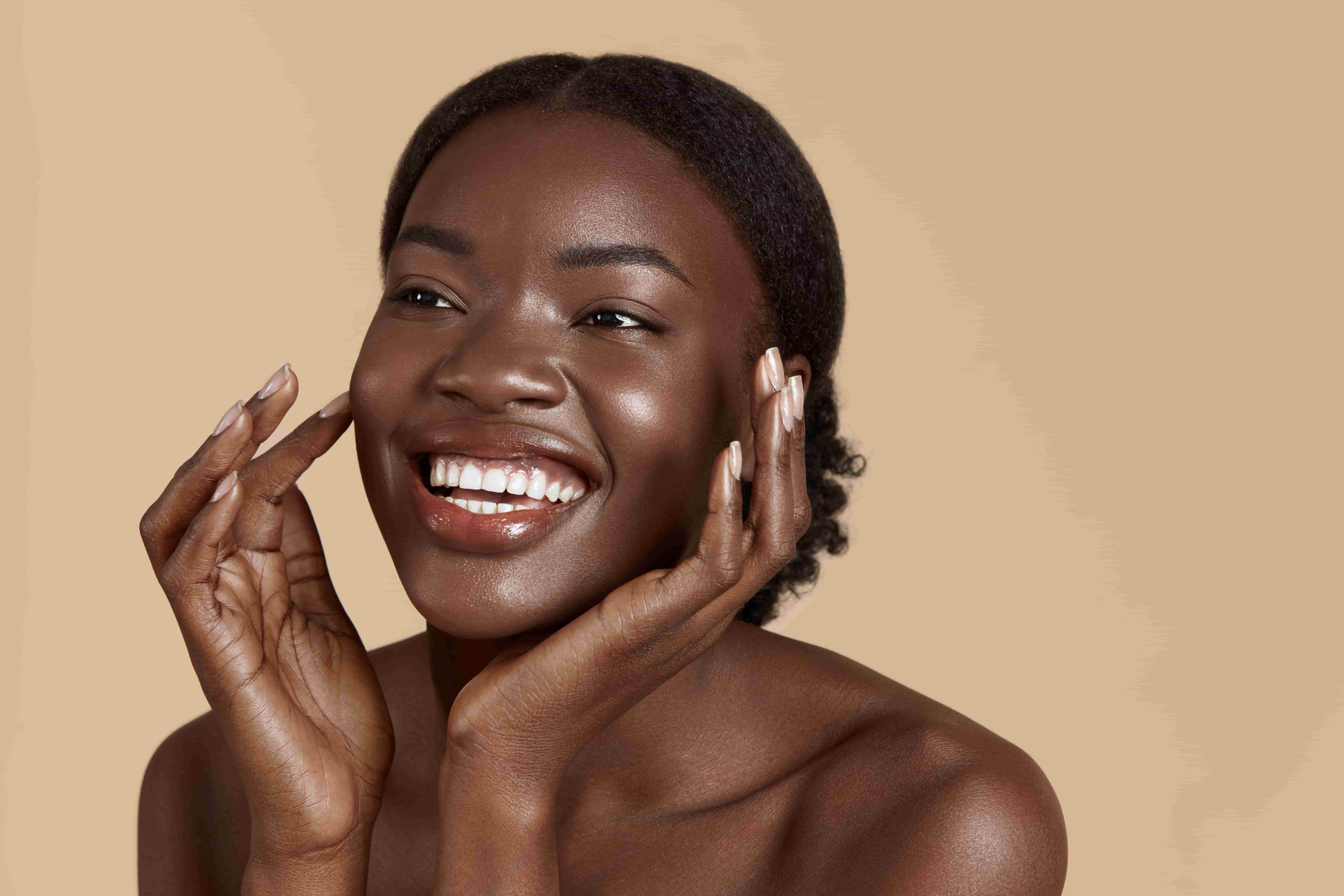 Mulher negra sorrindo com a pele radiante e bem cuidada, tocando o rosto com as mãos, em fundo neutro bege.