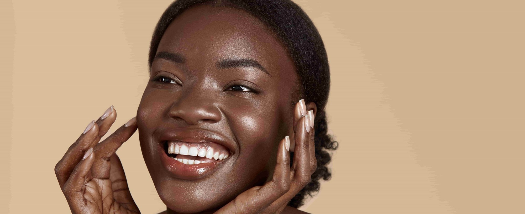 Mulher negra sorrindo com a pele radiante e bem cuidada, tocando o rosto com as mãos, em fundo neutro bege.
