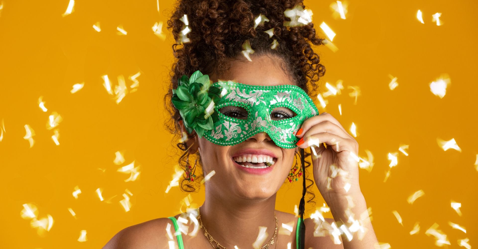 Close-up de uma pessoa sorridente com cabelo crespo escuro, levantando parcialmente uma máscara de carnaval verde e prateada.