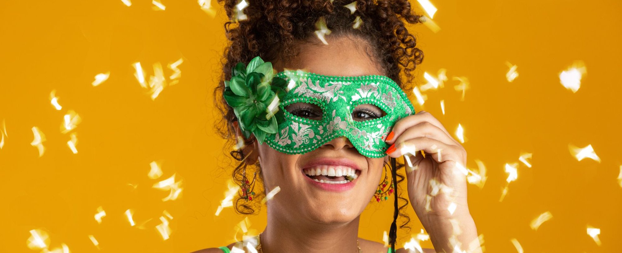 Close-up de uma pessoa sorridente com cabelo crespo escuro, levantando parcialmente uma máscara de carnaval verde e prateada.