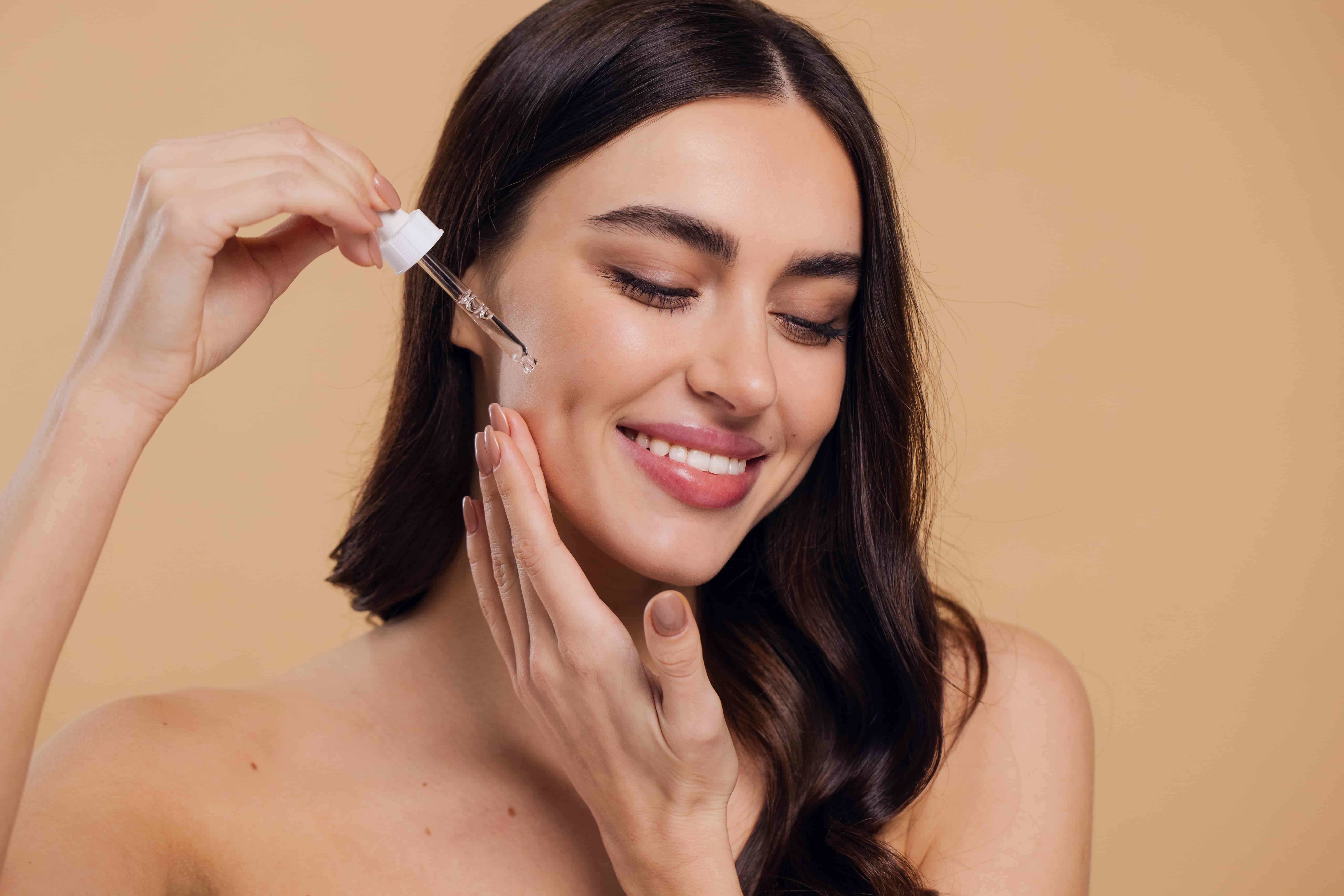 Uma jovem sorri ao aplicar um sérum hidratante em sua pele facial, enquanto toca o rosto levemente.
