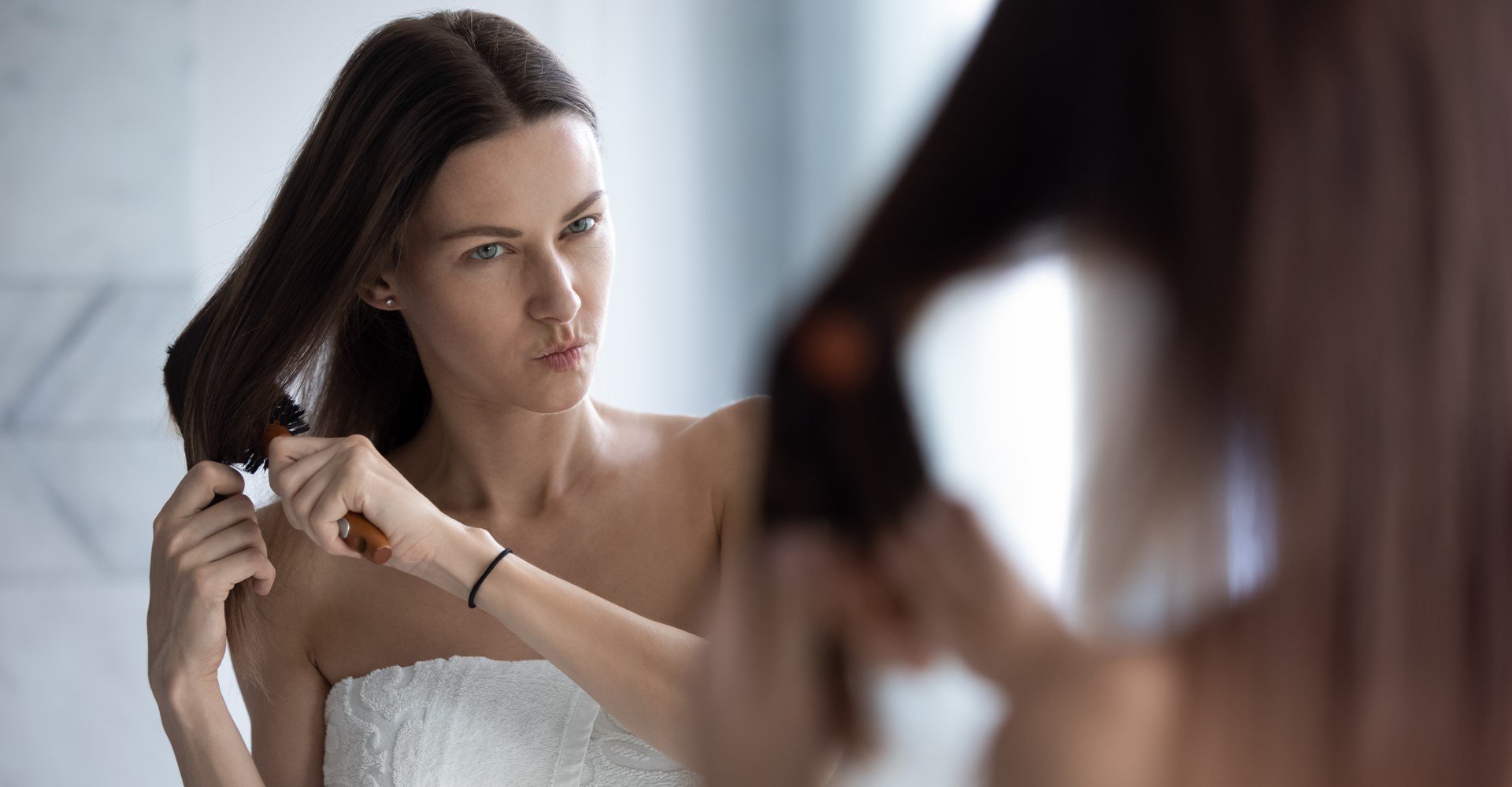 Modelo branca se olhando no espelho passando escova em seus cabelos