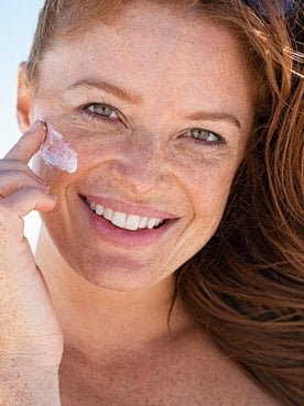 Modelo ruiva passando protetor solar no rosto e sorrindo