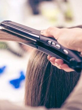 Modelo branca se olhando no espelho e passando chapinha no cabelo