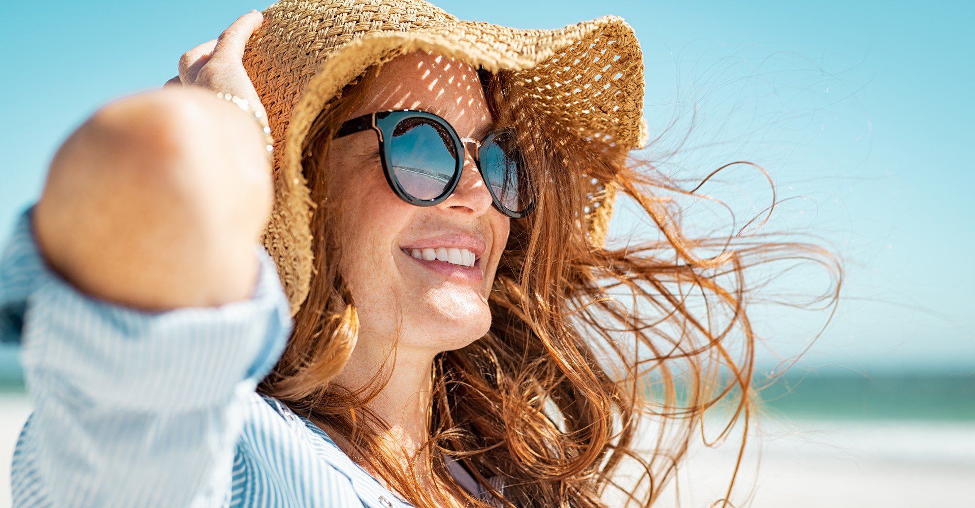 Modelo branca sorrindo com óculos de sol e chapéu cor palha