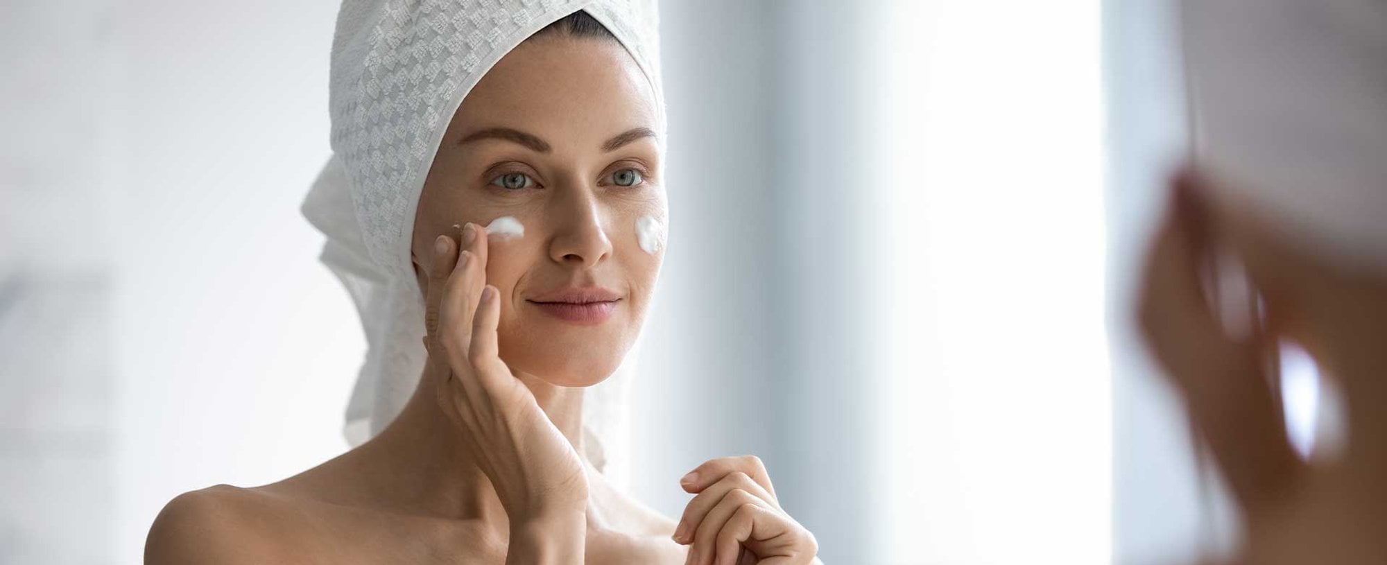 Mulher com toalha na cabeca e passando produto no rosto