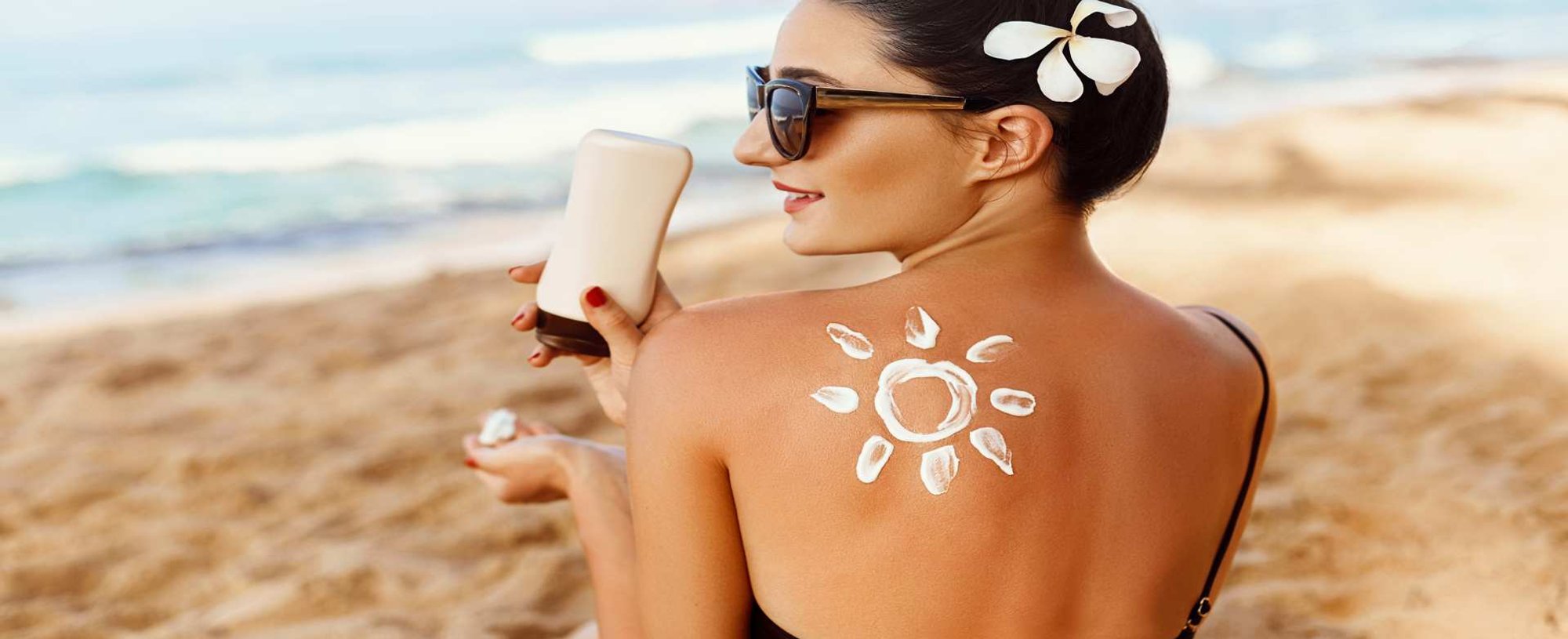 Modelo branca, na praia, sorrindo e passando protetor solar na mão