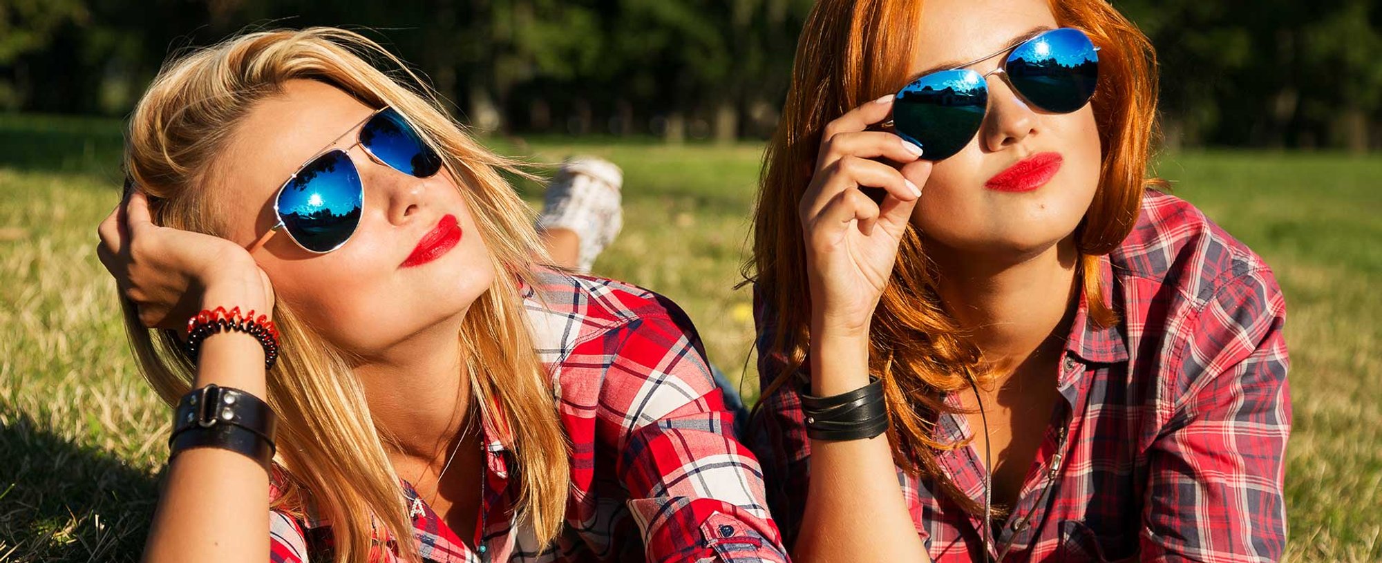 Duas mulheres brancas de óculos de sol deitadas na grama. Uma loira e a outra ruiva