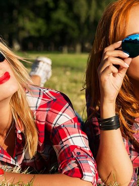 Duas mulheres brancas de óculos de sol deitadas na grama. Uma loira e a outra ruiva