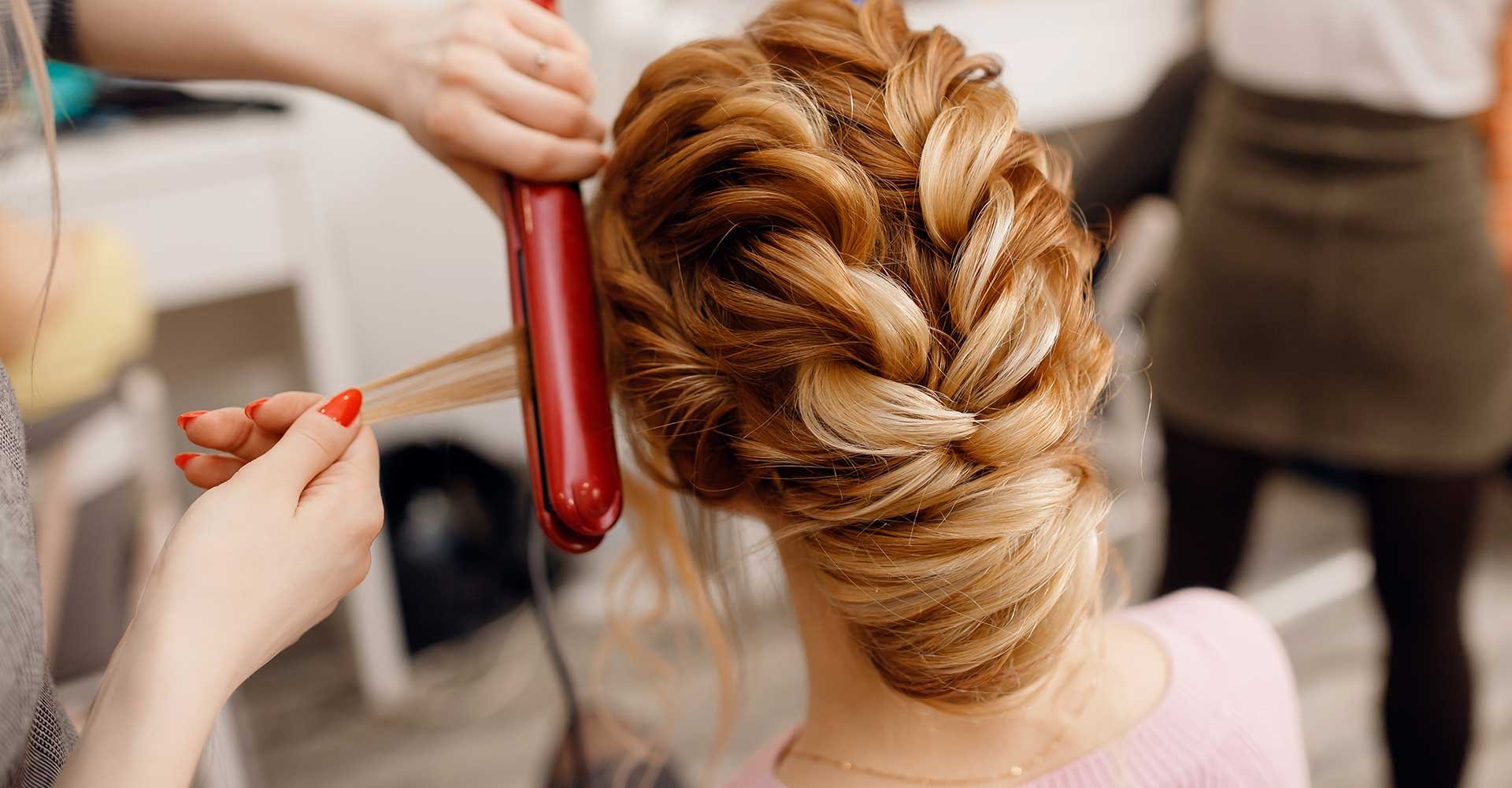 Imagem de uma mulher branca de costas e um profissional passando chapinha no seu cabelo ruivo com tranças 