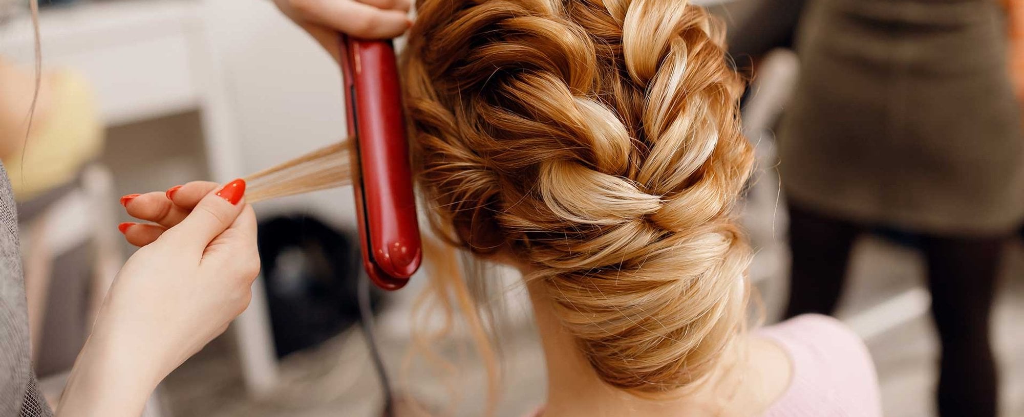 Imagem de uma mulher branca de costas e um profissional passando chapinha no seu cabelo ruivo com tranças