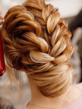Imagem de uma mulher branca de costas e um profissional passando chapinha no seu cabelo ruivo com tranças
