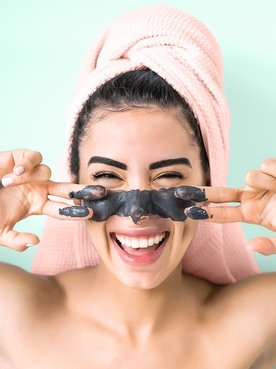 Imagem de mulher com toalha na cabeça e com mascara de argila preta no rosto.