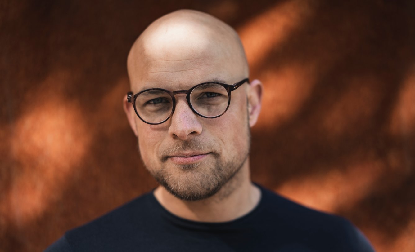 Imagem mostra um homem bonito e careca, usando óculos.