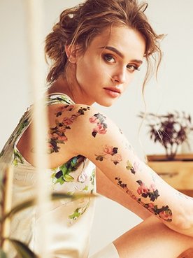 Imagem mostra uma mulher loira, sentada e com o braço tatuado.