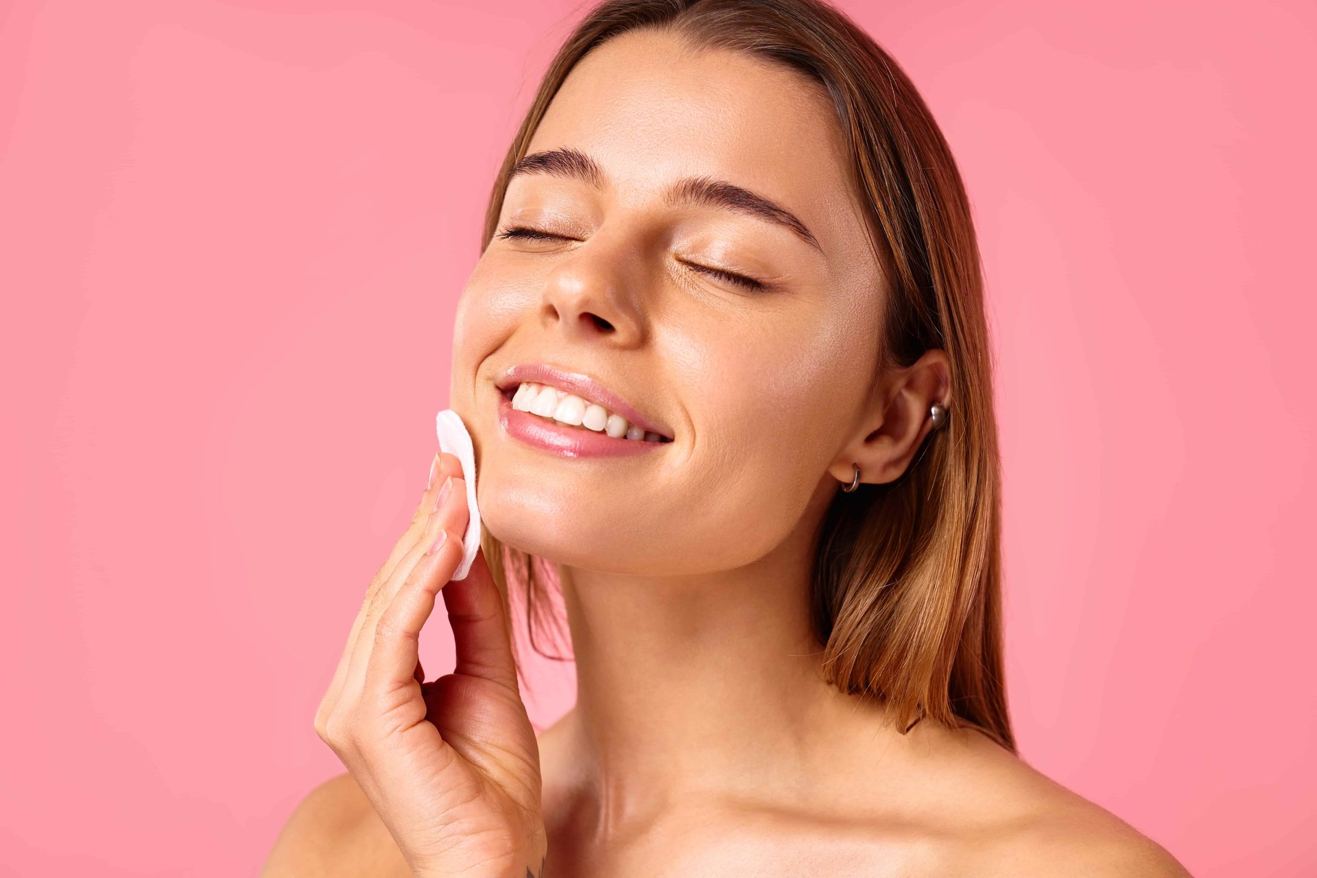Mulher sorrindo com os olhos fechados enquanto aplica água micelar no rosto com algodão, em um fundo rosa, transmitindo leveza e cuidado com a pele.