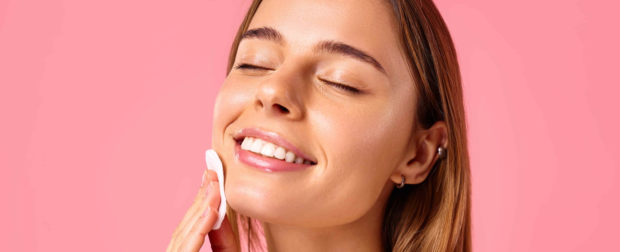 Mulher sorrindo com os olhos fechados enquanto aplica água micelar no rosto com algodão, em um fundo rosa, transmitindo leveza e cuidado com a pele.
