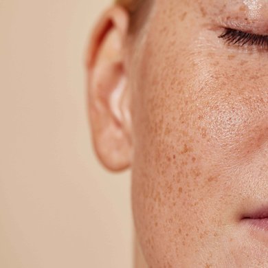 Rosto de pessoa com pele clara e sardas, olhos fechados, em close, com pele hidratada e acabamento natural sobre fundo bege.