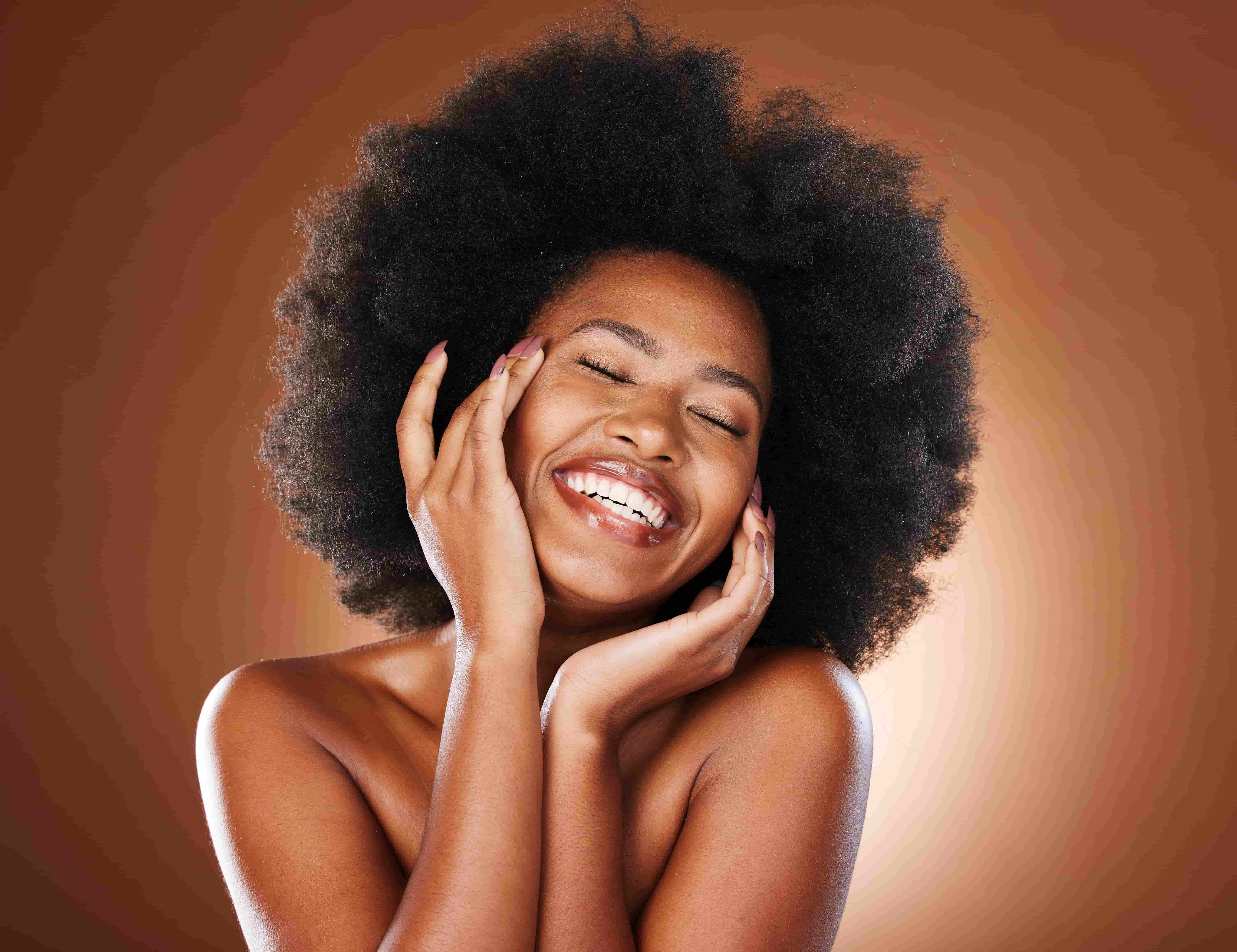 Mulher sorrindo com as mãos no rosto, exibindo pele radiante e hidratada, com cabelo afro natural em fundo marrom degradê.