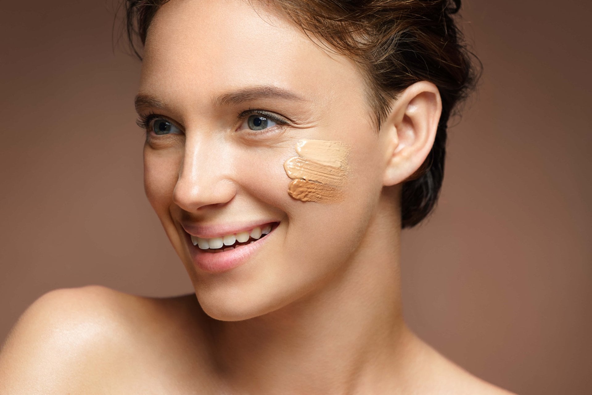 Mulher sorrindo com amostras de base em dois tons aplicados na bochecha para encontrar o tom ideal para a maquiagem do dia a dia.